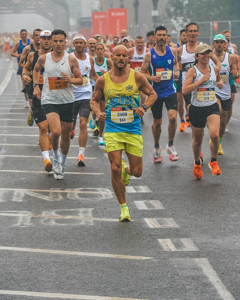 Man wearing yellow running attire runs The London Marathon, he leads a pack of runners. Man is Simon Radcliffe from the Radcliffe Group.